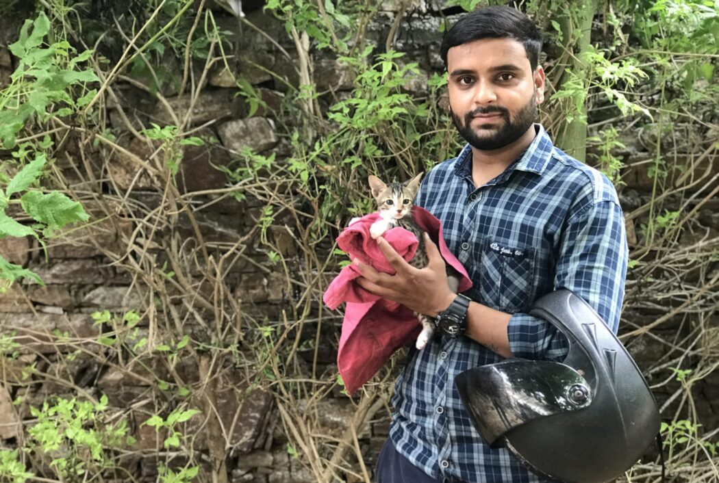 An Indian man in a blue chequered shirt is holding the new tiny tabby kitten he has adopted. His left arm is looped through his black motorcycle helmet and the kitten is wrapped in a pink blanket. Behind them is a stone wall and green bushes.