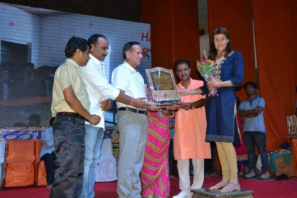 Rachel is wearing a dark blue salwar and cream leggings. She has a bunch of flowers in one hand and is touching an award with the other hand. 5 more people are present – 4 males and 1 female but she is mostly hidden, wearing a pink sari. The man standing nearest Rachel is wearing an orange shirt and white trousers.