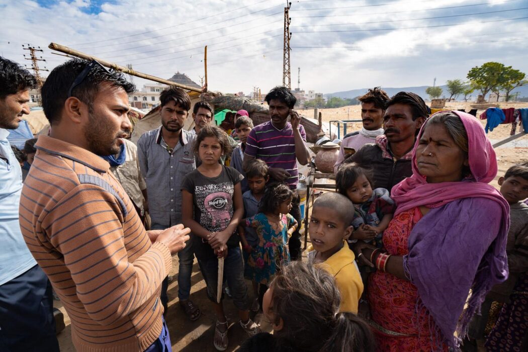 A group of around sixteen people are standing together, centred around a man on the left who is speaking and gesturing at them.