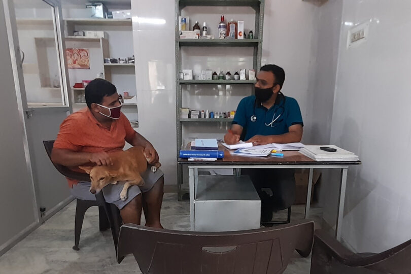 Male Veterinary Doctor wearing a dark green T shirt and stethoscope is sitting at a desk talking to a man on his right hand side who is wearing an orange T shirt and grey shorts, that has a tan dog that is sitting on his knee. Both are wearing masks. In the background there is a shelf with medicines on it.