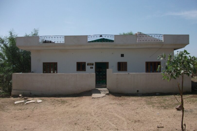 The forefront of the photo is a sandy area. Alter this is a large single story building painted white with a main double gate and wooden windows either side. There is a small wall making a veranda area. There are decorative black railings around the roof space.