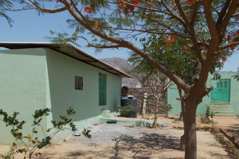 In the forefront is a sanded area with small trees growing. Behind this area is a single story green painted building with steps front and centre leading to double metal gates.