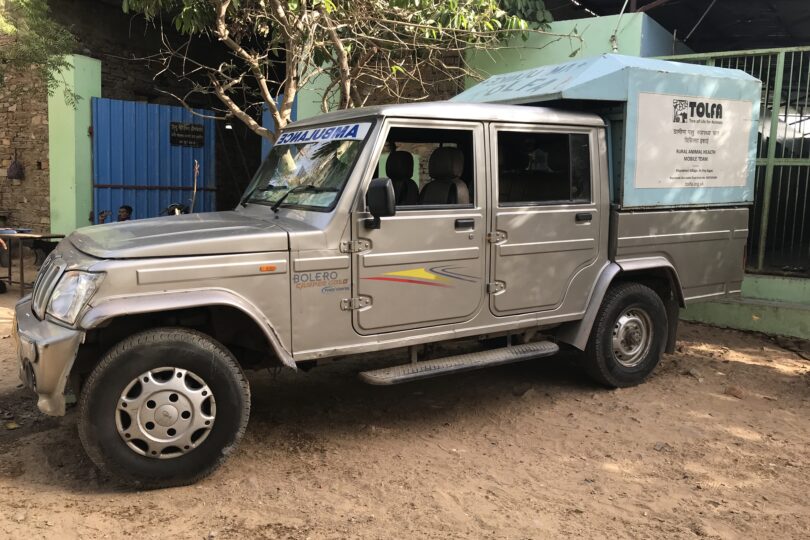 A silver vehicle with a light blue cab is backed up to a green gate. There is a big blue gate attached to a green pillar in the background.