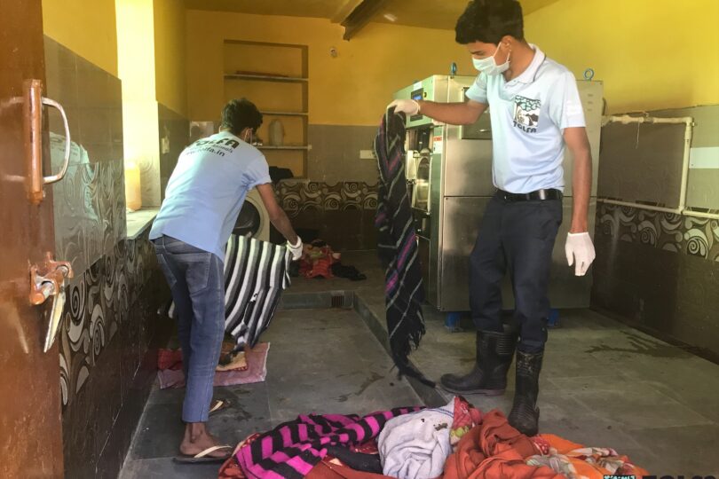 Two members of staff are sorting out multi coloured blankets on a stone floor. Behind them to the right is an industrial stainless steel washing machine.