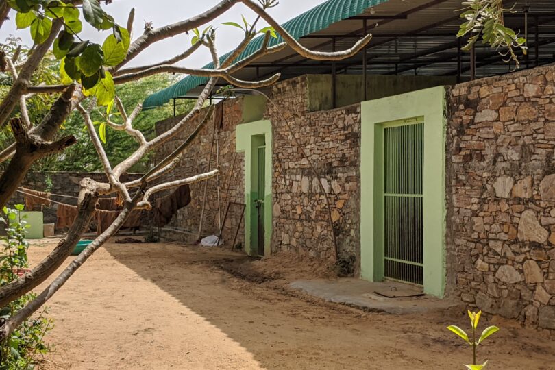 In the forefront is a sanded area with small trees growing. Behind this area is a single story green painted building with steps front and centre leading to double metal gates.