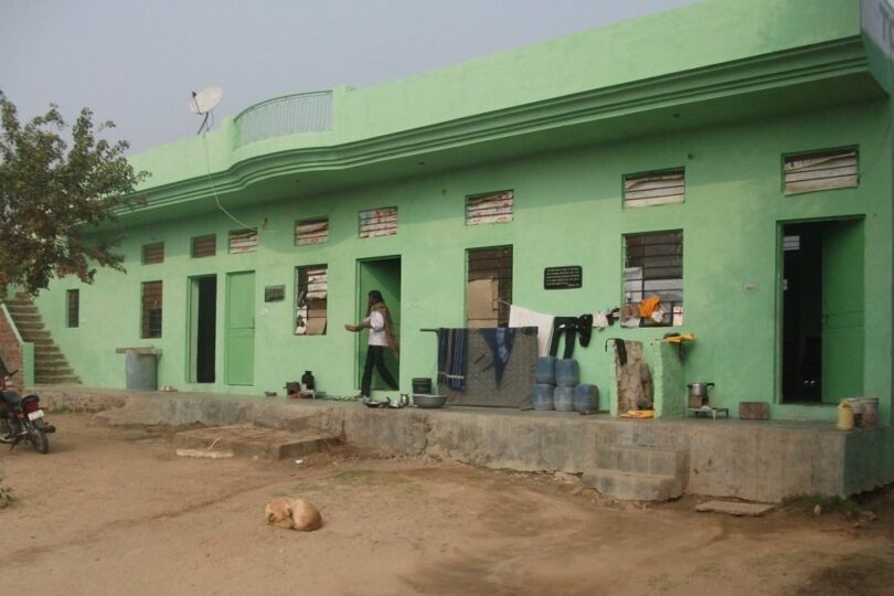 Photo shows long, low building painted green on the right hand side. There are 4 individual doors leading into houses. Washing is hanging outside the houses and a man is walking carrying a bowl.