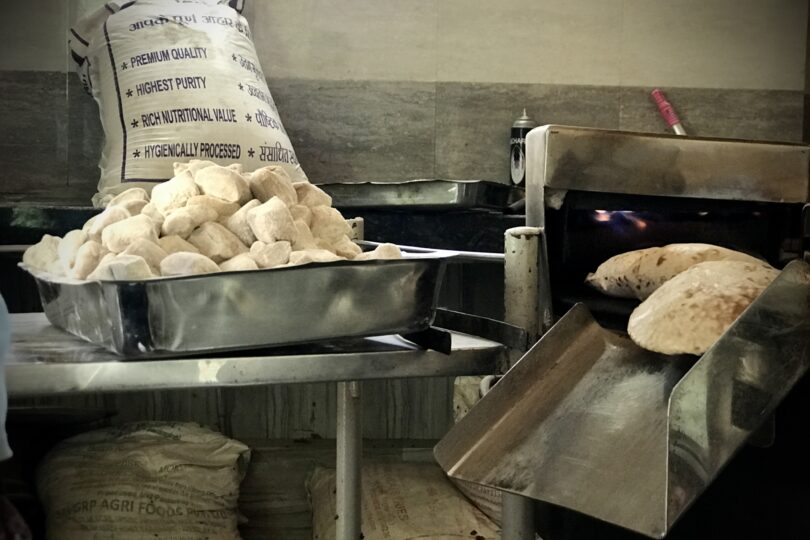 View of the roti (chapatti) making machine in action. Freshly cooked round rotis are coming out of the fire and down the steel chute and there is a large tray of dough balls and a bag of flour on the table next to the machine.