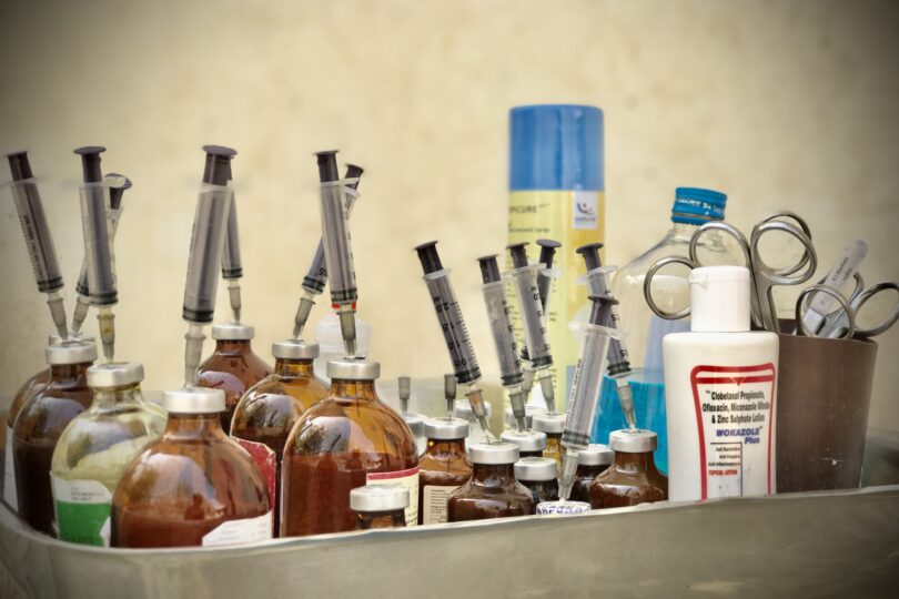 A deep steel tray filled with brown glass medicine injection bottles. Each bottle has a needle and syringe pierced downwards into it. There is also a pot of metal scissors, a bottle of dog shampoo and a can of wound spray.