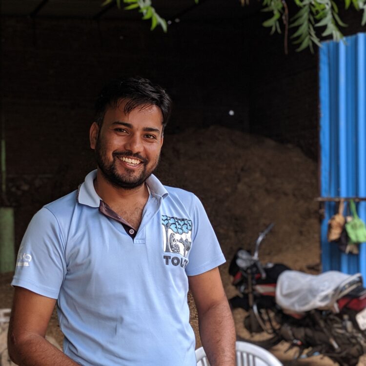 A TOLFA staff wearing a pale blue polo shirt if facing the camera and smiling. There is dry fodder and motorbikes in the background.